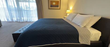 Una cama bien hecha con un edredón azul oscuro y almohadas blancas, una lámpara de noche y un cuadro en la pared.