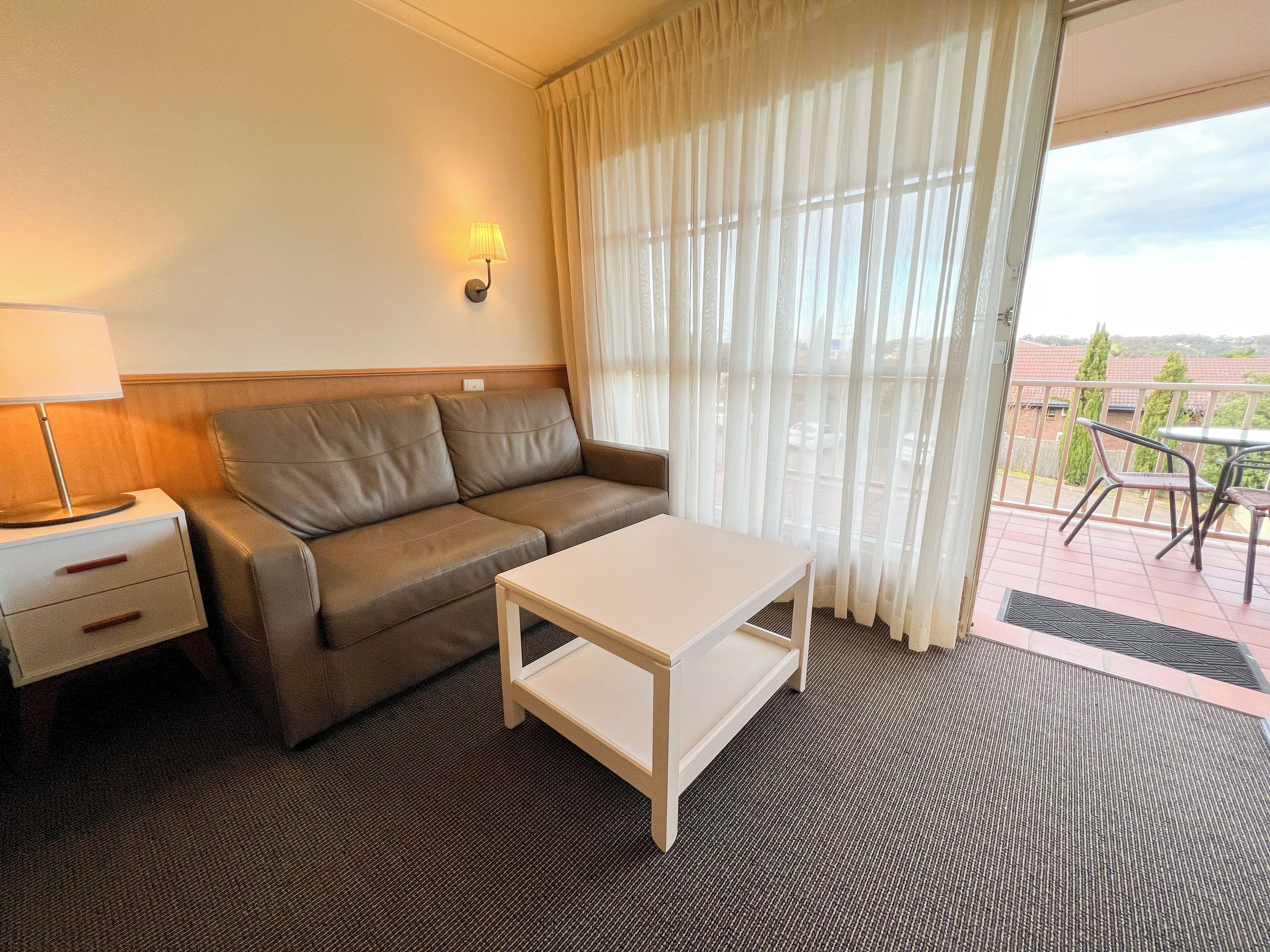 Habitación de hotel con sofá, una mesita, lámpara y vistas al exterior a través de cortinas translúcidas.