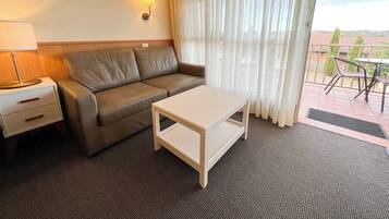 Habitación de hotel con sofá, una mesita, lámpara y vistas al exterior a través de cortinas translúcidas.
