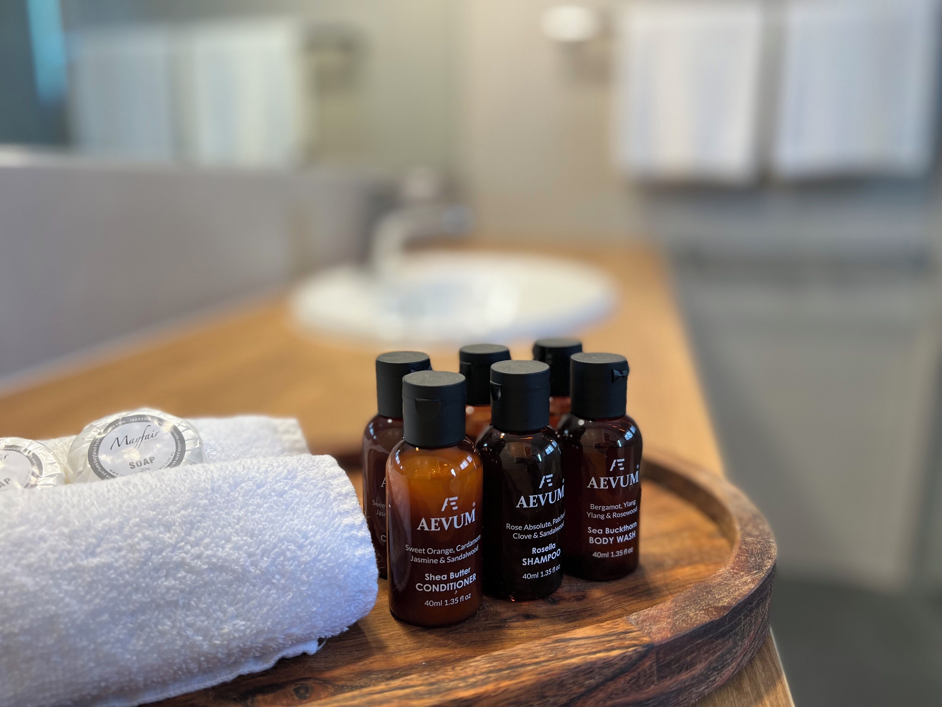 A wooden tray with bottles of AEVUM body wash and shampoo, a white towel, and a soap bottle on a bathroom counter.