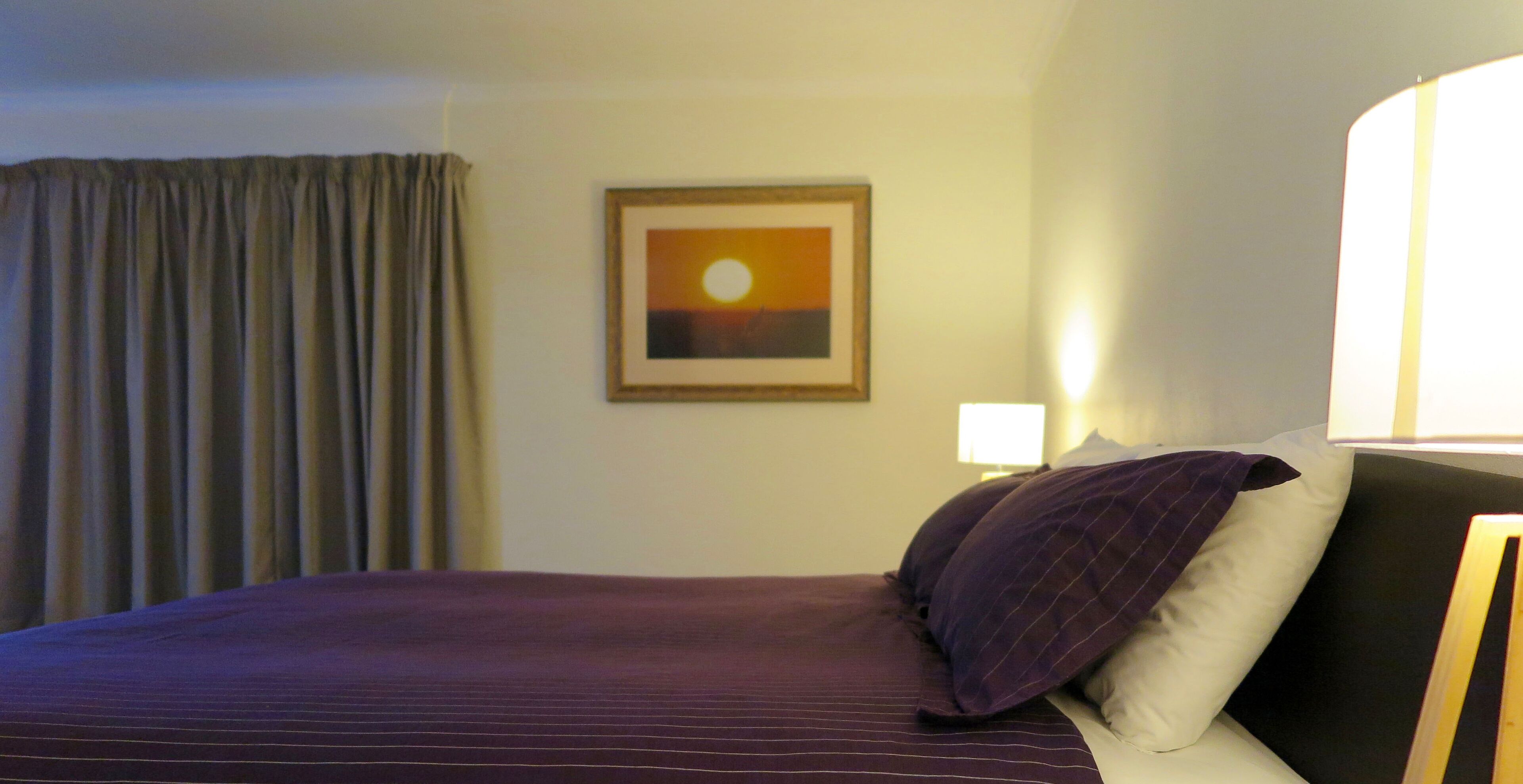 Habitación de hotel con cama, un cuadro enmarcado de un atardecer, una lámpara y una ventana con cortinas.
