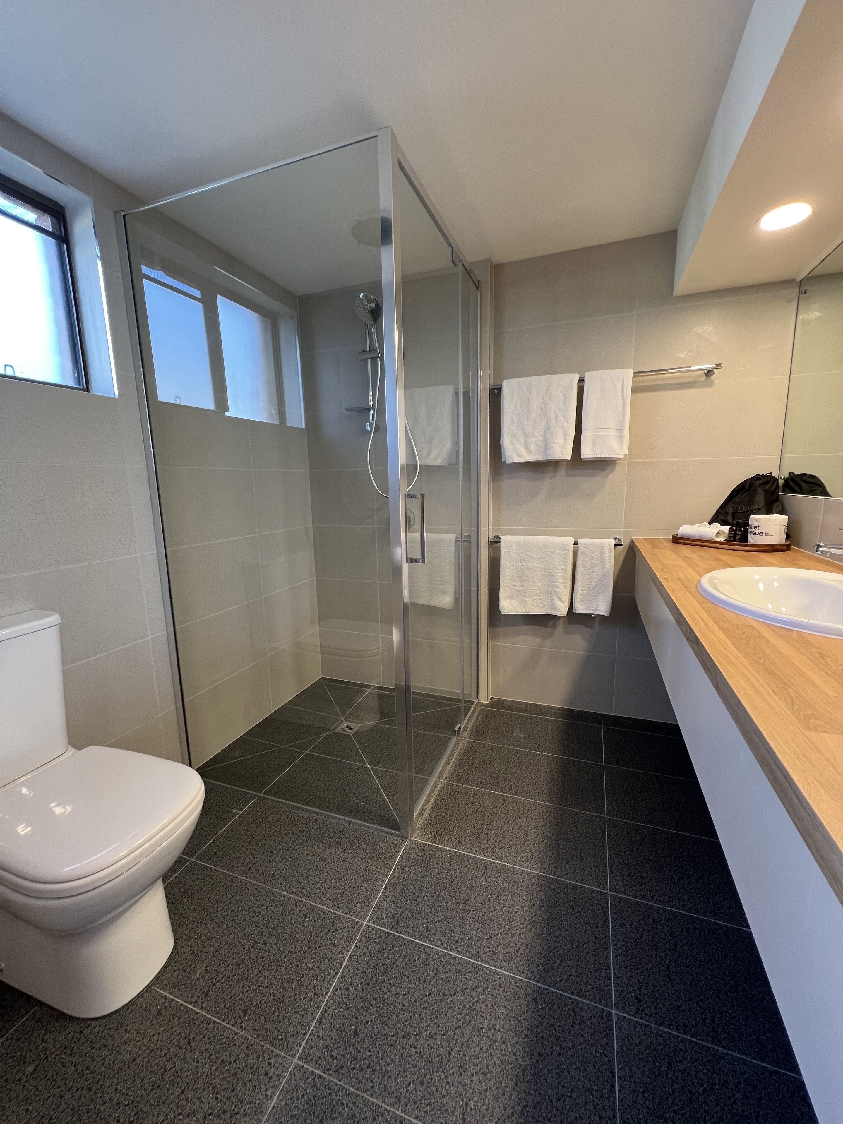 Une salle de bain moderne dotée d’une douche à parois de verre, de toilettes, d’un lavabo avec un plan de travail en bois et d’un porte-serviettes avec plusieurs serviettes.