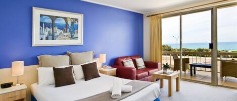 A hotel room with a large bed, a red sofa, a small table, and a balcony with a view of the sea.