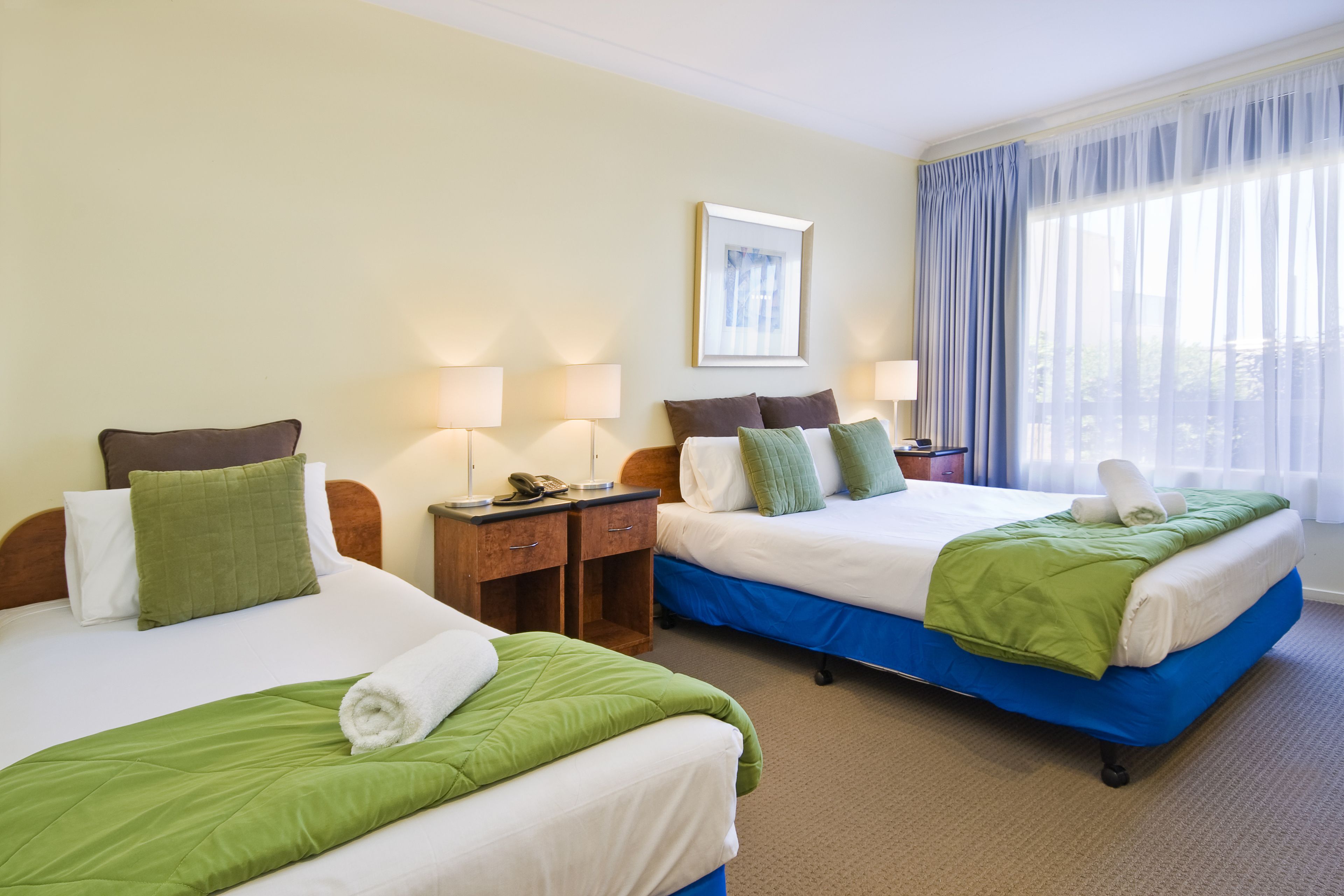 Duas camas de solteiro com roupa de cama verde e branca, mesas de cabeceira de madeira e uma janela grande com cortinas.