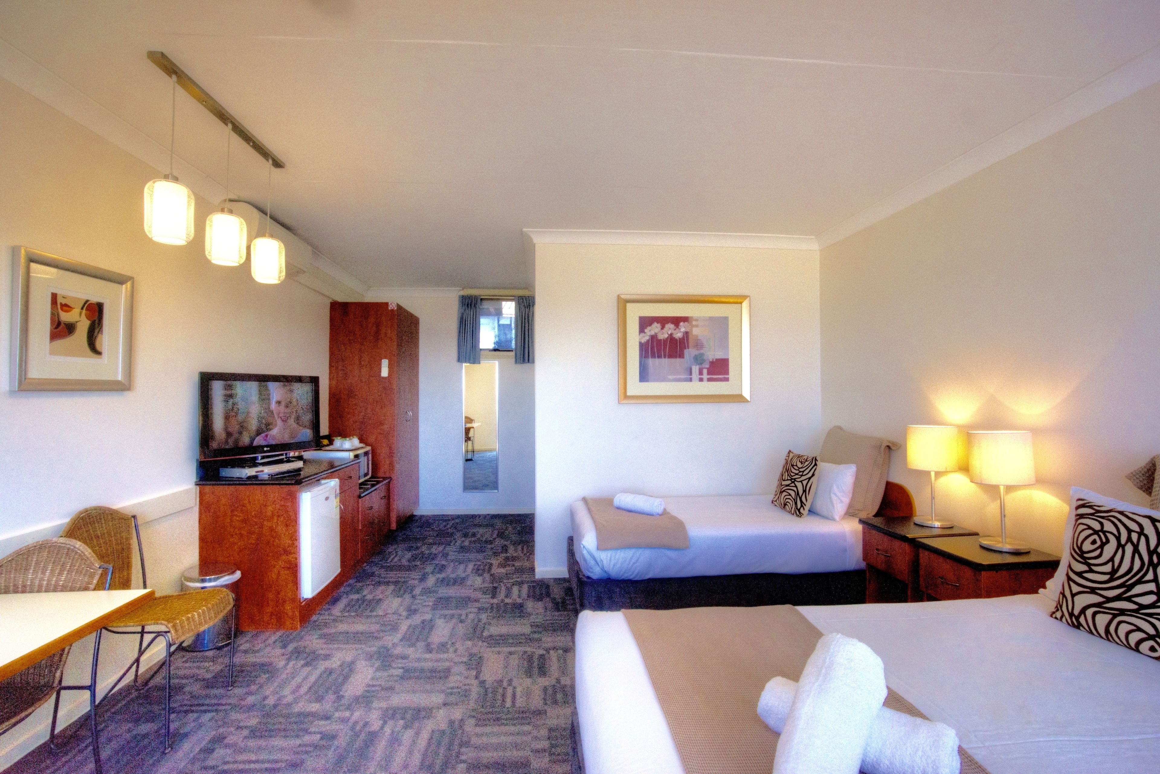 Habitación de hotel con dos camas, un escritorio con televisor, una kitchenette pequeña y un baño visible a través de una puerta abierta.