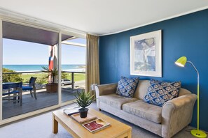 A living room with a beige sofa, a wooden coffee table, and a view of the ocean through large sliding glass doors. - Sandridge Motel (Lorne)
