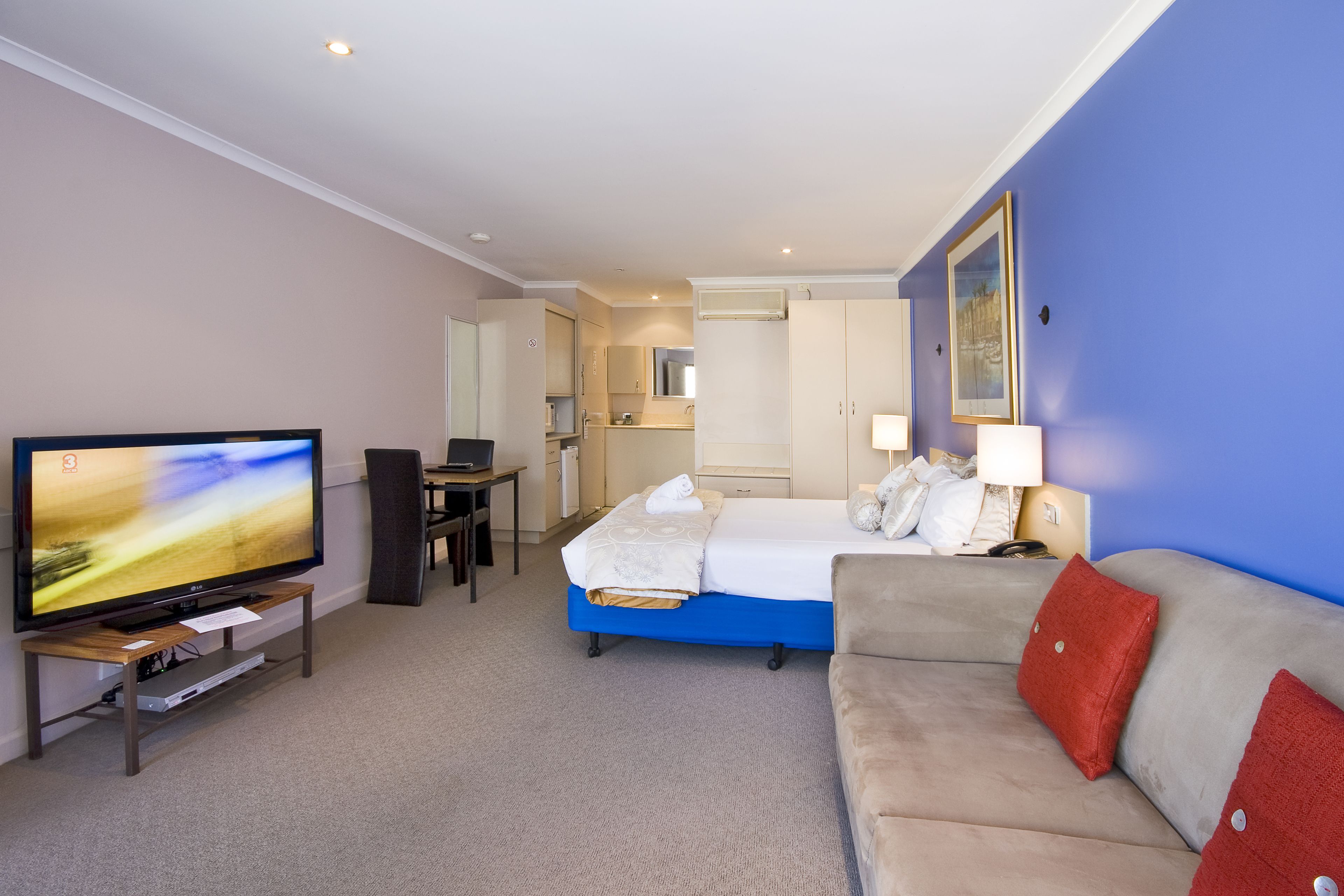 A hotel room with a bed, a sofa, a TV, a desk, and a bathroom visible in the background.