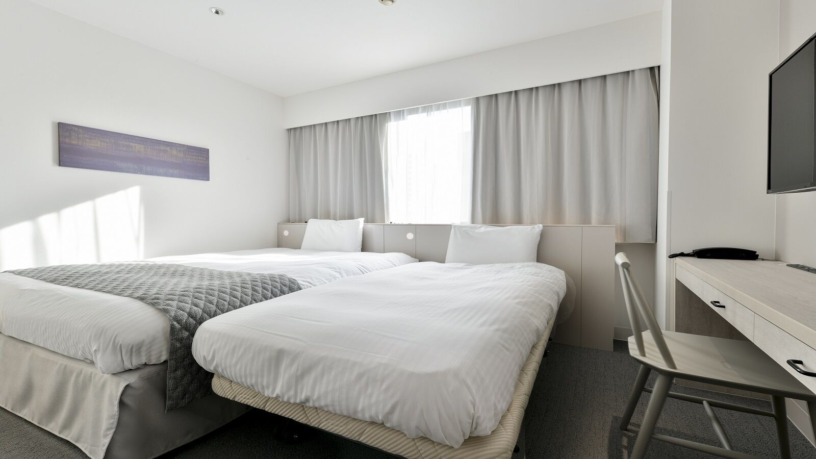 Ein Hotelzimmer mit zwei Betten, einem Schreibtisch, einem Stuhl, einem Fernseher und einem Fenster mit Vorhängen.