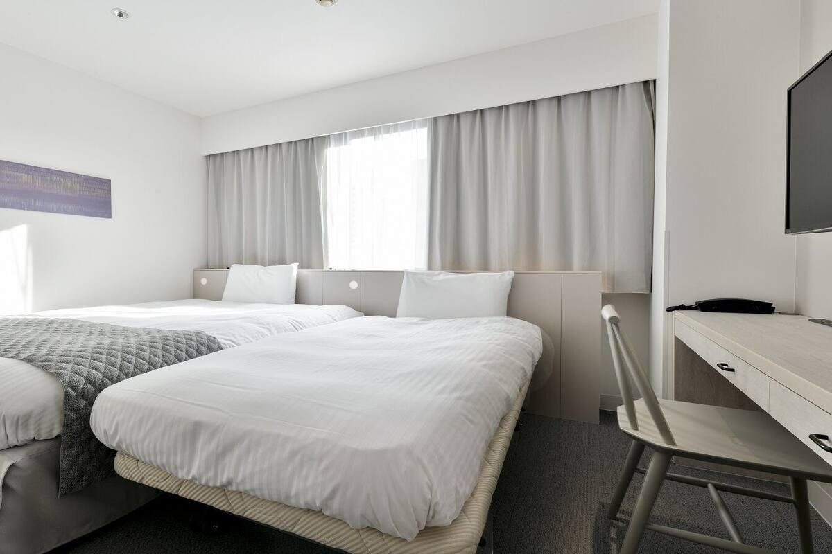 A hotel room with two beds, a desk, a chair, a TV, and a window with curtains.