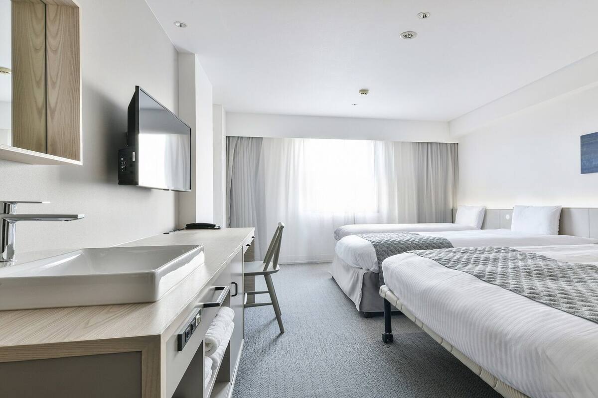 A hotel room with a desk, two beds, a TV, and a large window with curtains.