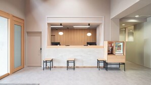 Un área de recepción con un mostrador de madera, dos monitores de computadora y un refrigerador pequeño. Hay tres sillas de madera frente al mostrador.