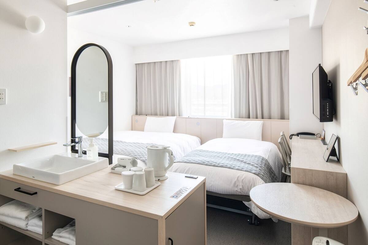 A hotel room with a large mirror, two beds, a bedside table with a kettle, a small round table, and a TV mounted on the wall.