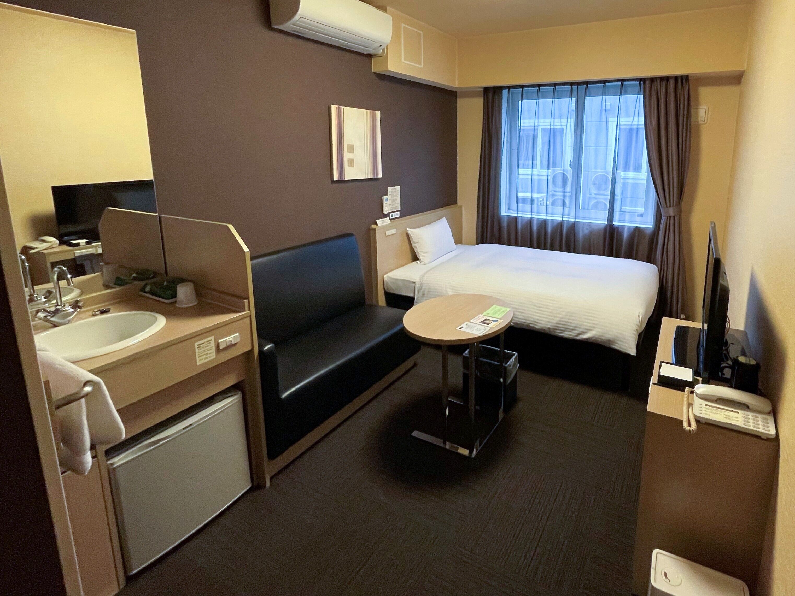 Ett hotellrum med en säng, ett litet bord, en soffa, ett skrivbord med handfat och en spegel.