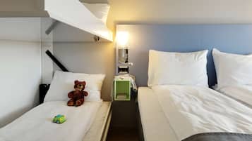 Habitación de hotel con dos camas, un osito de peluche, un coche de juguete y una mesita de noche con una lámpara.