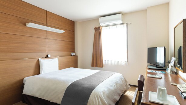 A hotel room with a bed, a desk with a TV, a mirror, and an air conditioning unit. - Vessel Hotel Higashi - Hiroshima (Higashihiroshima)