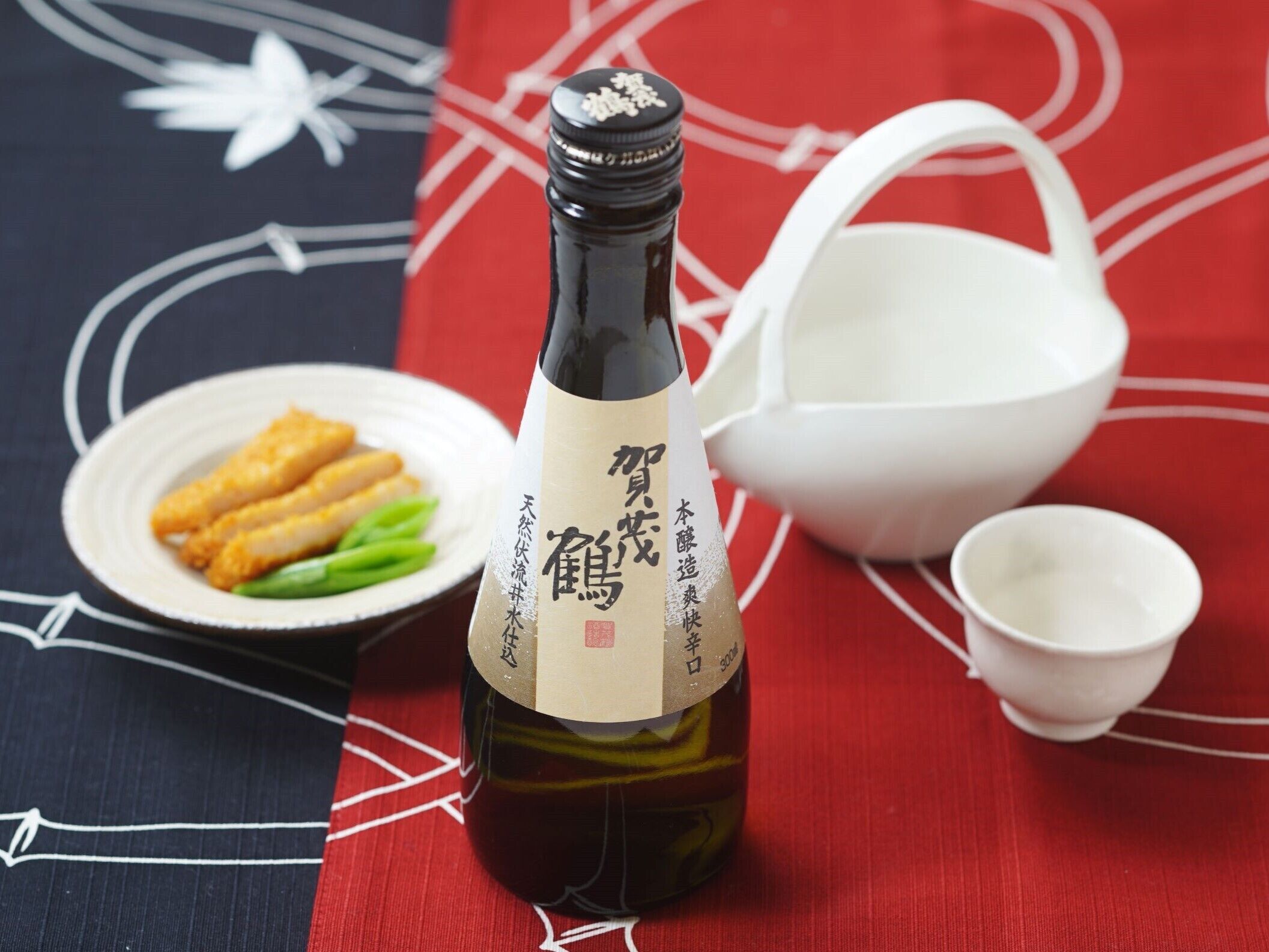 A bottle of sake with a label, a plate with food, and a white bowl on a tablecloth with a red and black pattern.