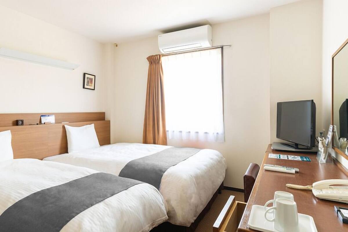 A hotel room with two beds, a desk with a computer and phone, and an air conditioning unit.