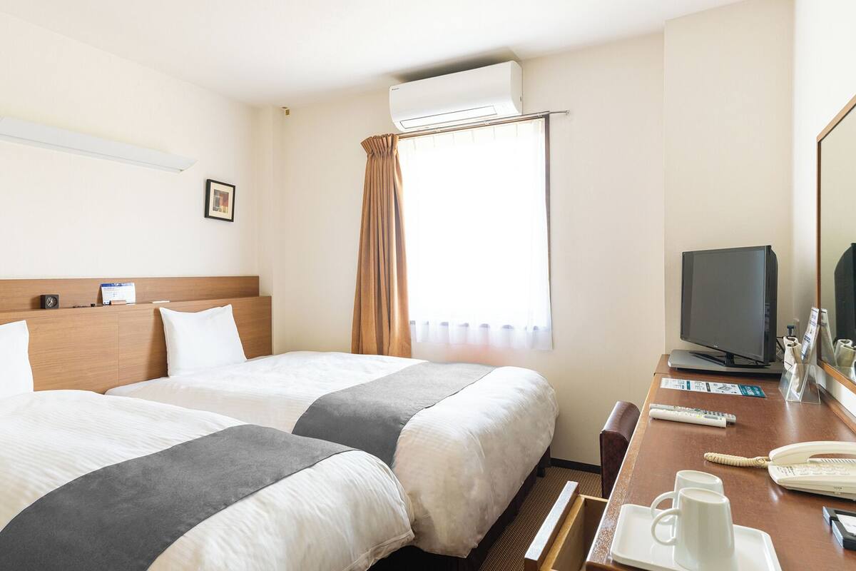 A hotel room with two beds, a desk with a computer and phone, an air conditioner, and a window with curtains.