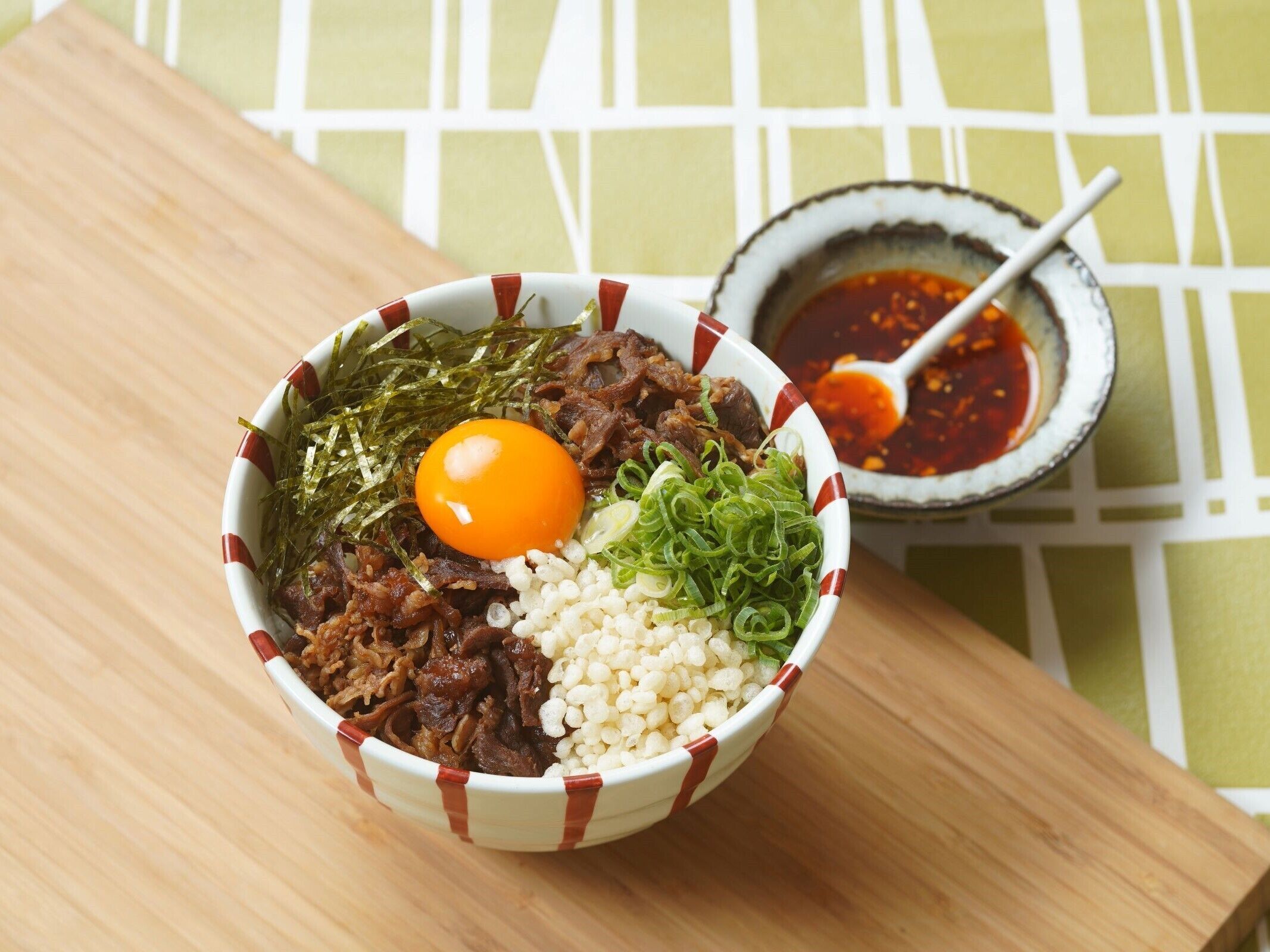 A bowl of food with various ingredients including meat, rice, and a boiled egg, accompanied by a small bowl of sauce.