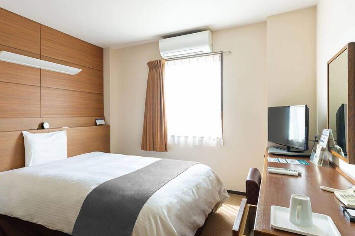 A hotel room with a bed, a desk with a TV, a mirror, and an air conditioning unit.