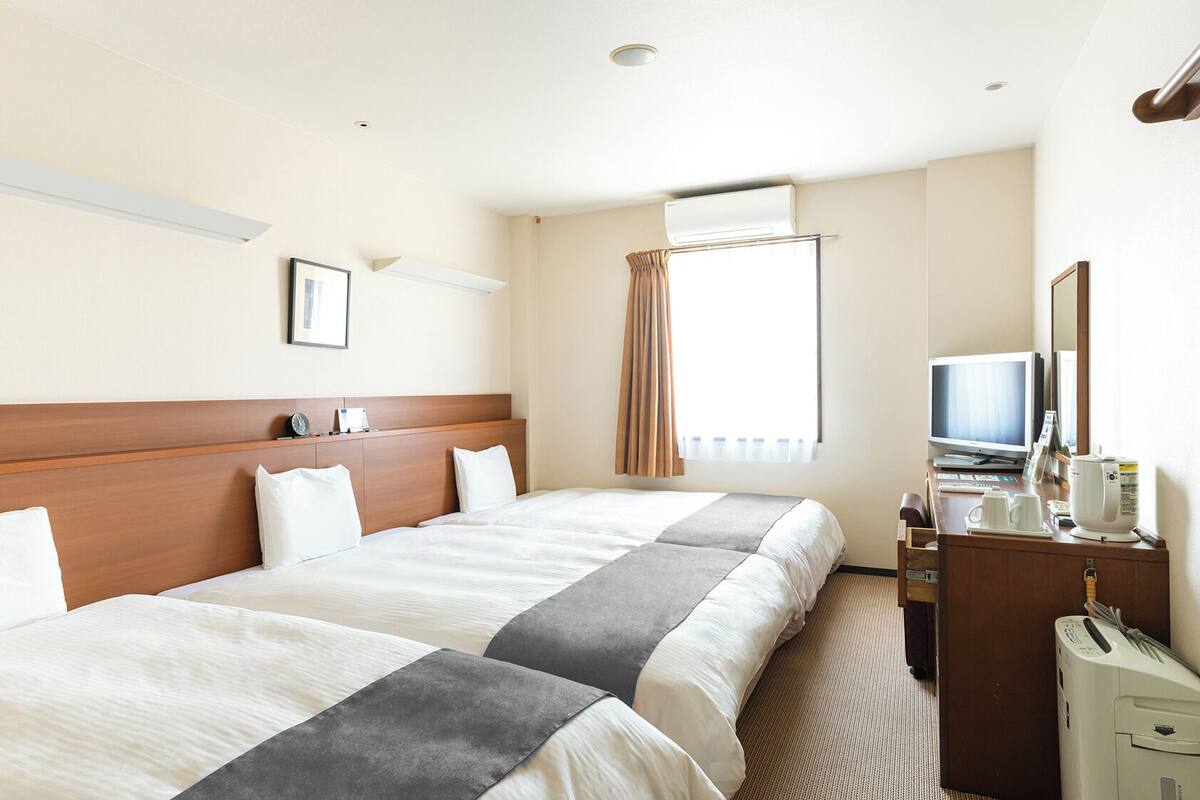 A hotel room with two beds, a desk with a TV, a coffee maker, and an air conditioning unit.