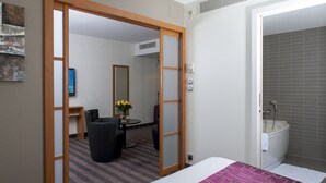 Habitación de hotel con cama, escritorio y silla, una mesita con flores, y baño con bañera y ducha.