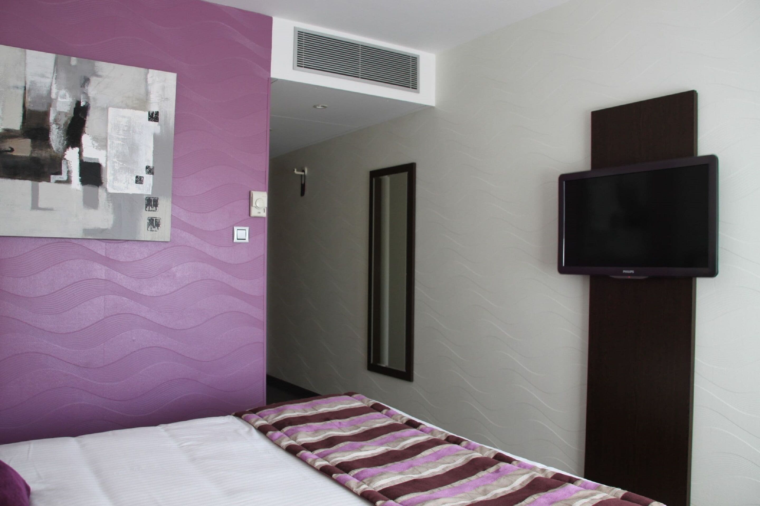 Habitación de hotel con cama, televisión en la pared, un espejo grande y un cuadro.