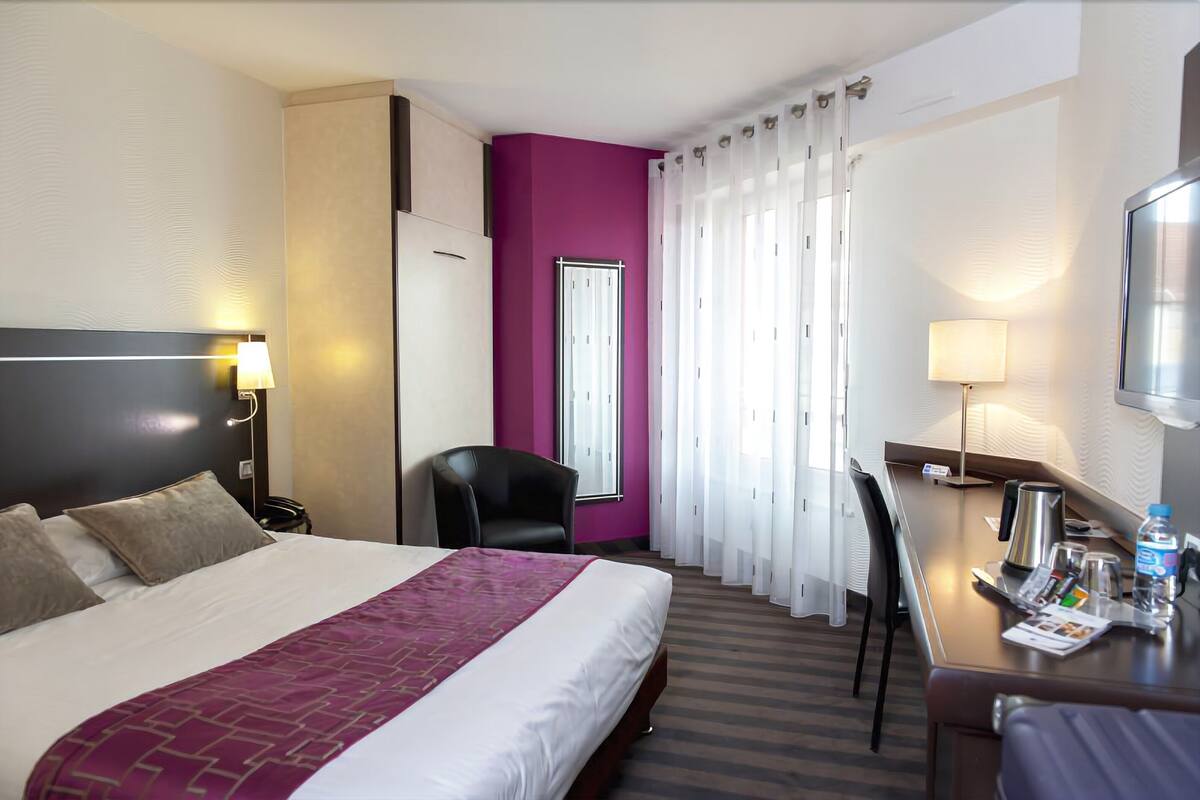 Habitación de hotel con cama, escritorio, silla, televisor y ventana con cortinas.