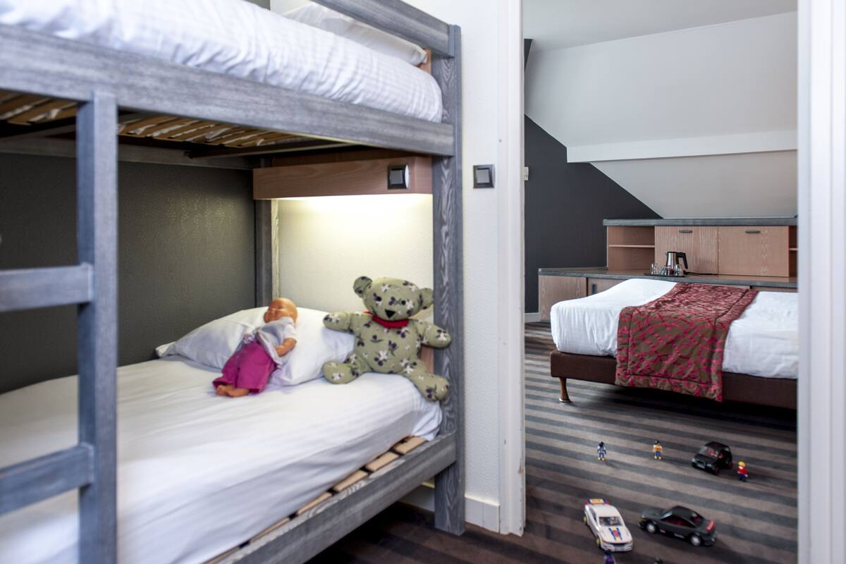 Habitación con literas, un oso de peluche en la cama inferior, una kitchenette pequeña y un coche de juguete en el suelo.