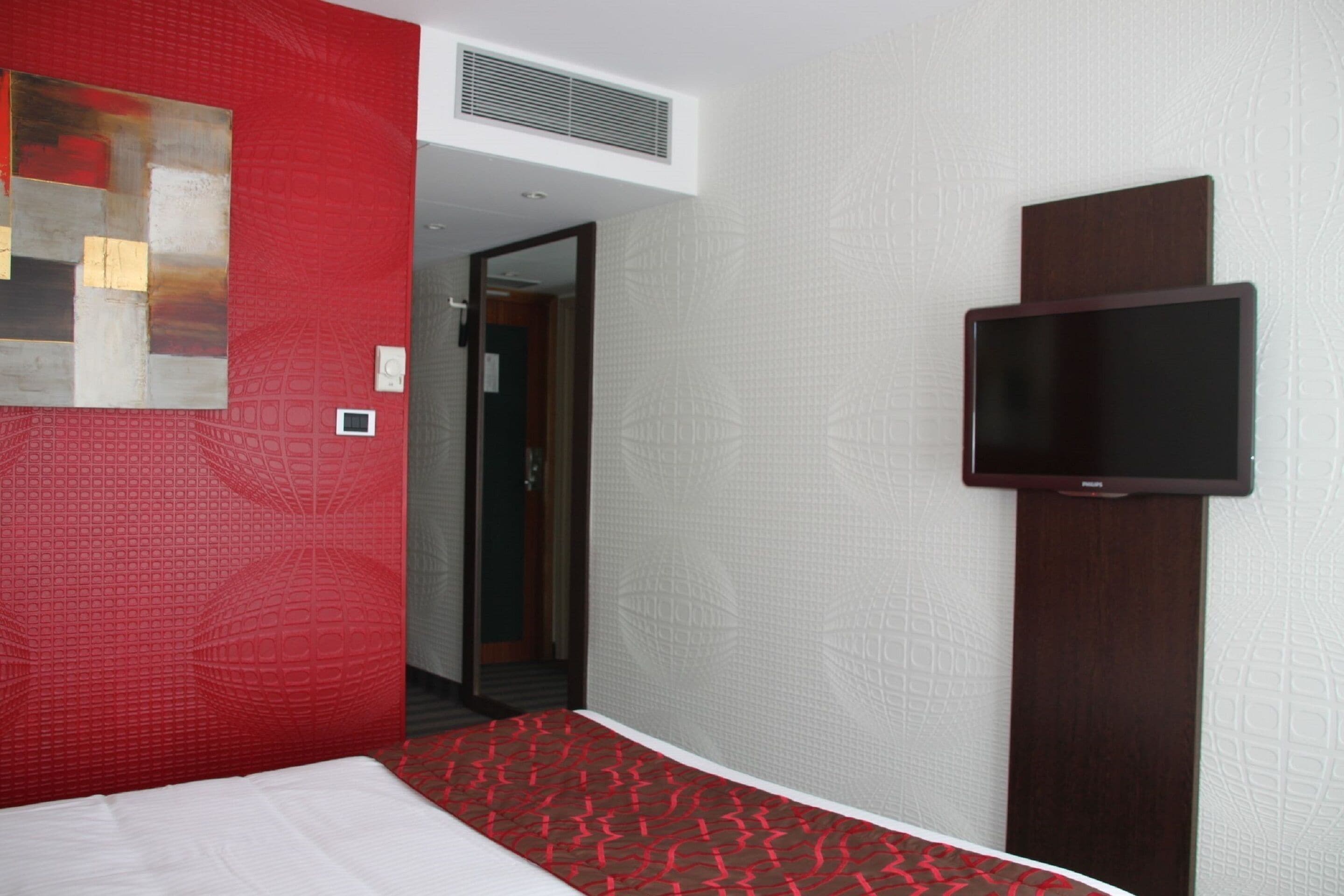 A hotel room with a bed, a television mounted on the wall, and a red accent wall.