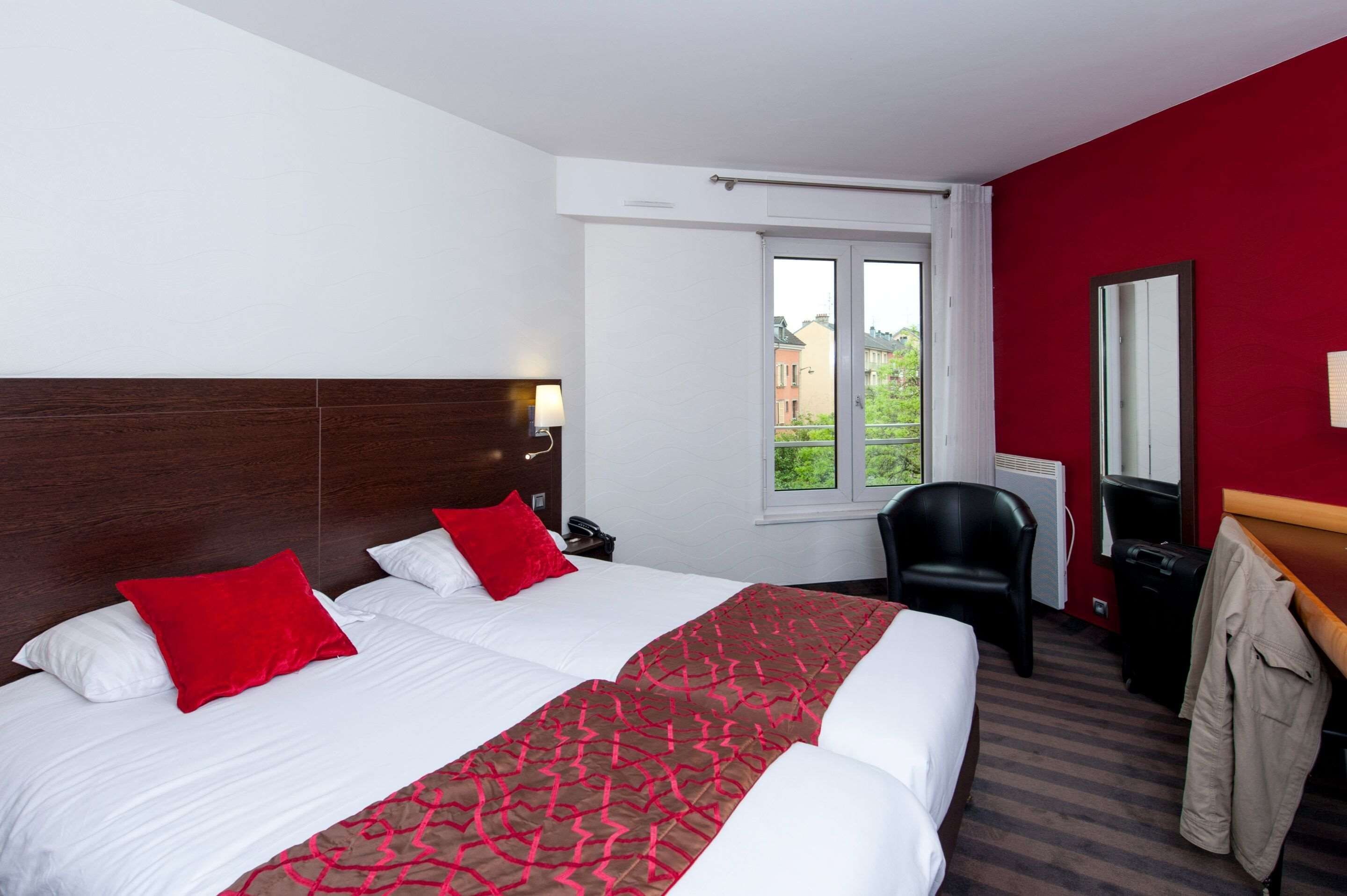 Habitación de hotel con dos camas, una pared de acento roja, una ventana con vistas, una silla y un perchero.