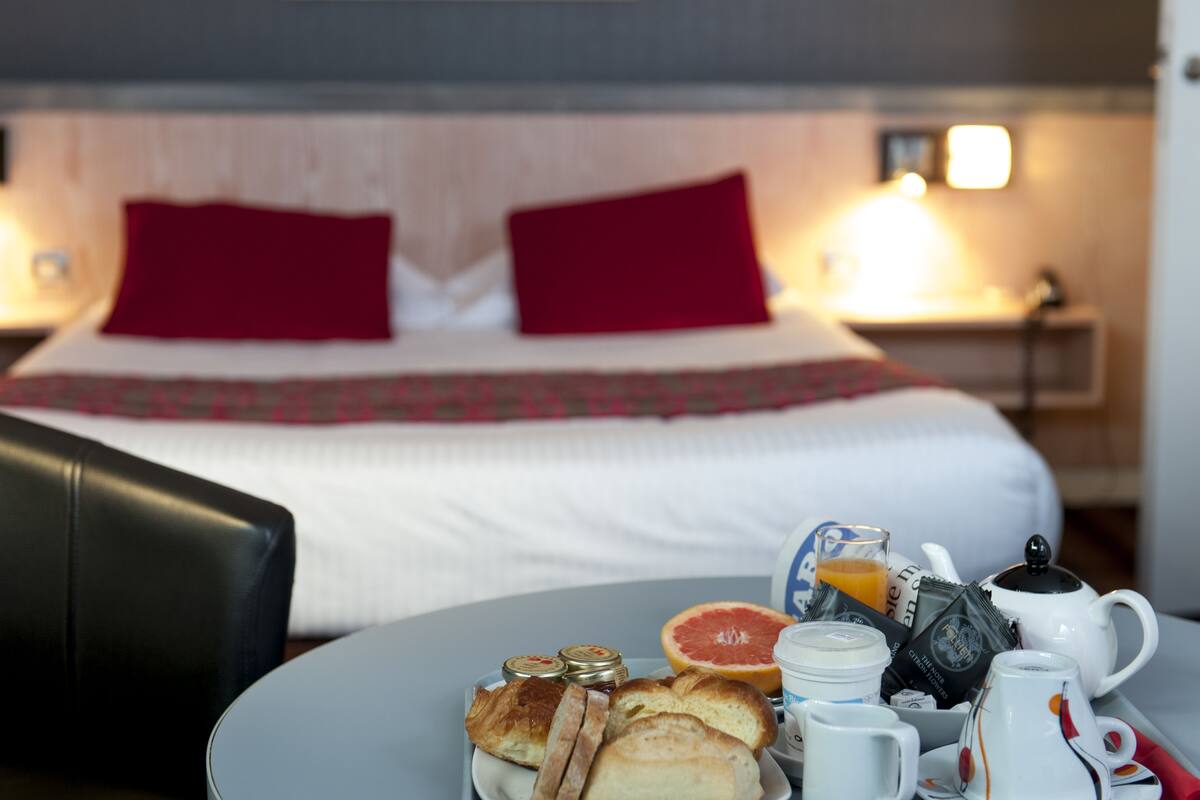 Una cama bien hecha con almohadas rojas, una mesita de noche con una lámpara y una bandeja de desayuno con pan, jugo y una tetera.