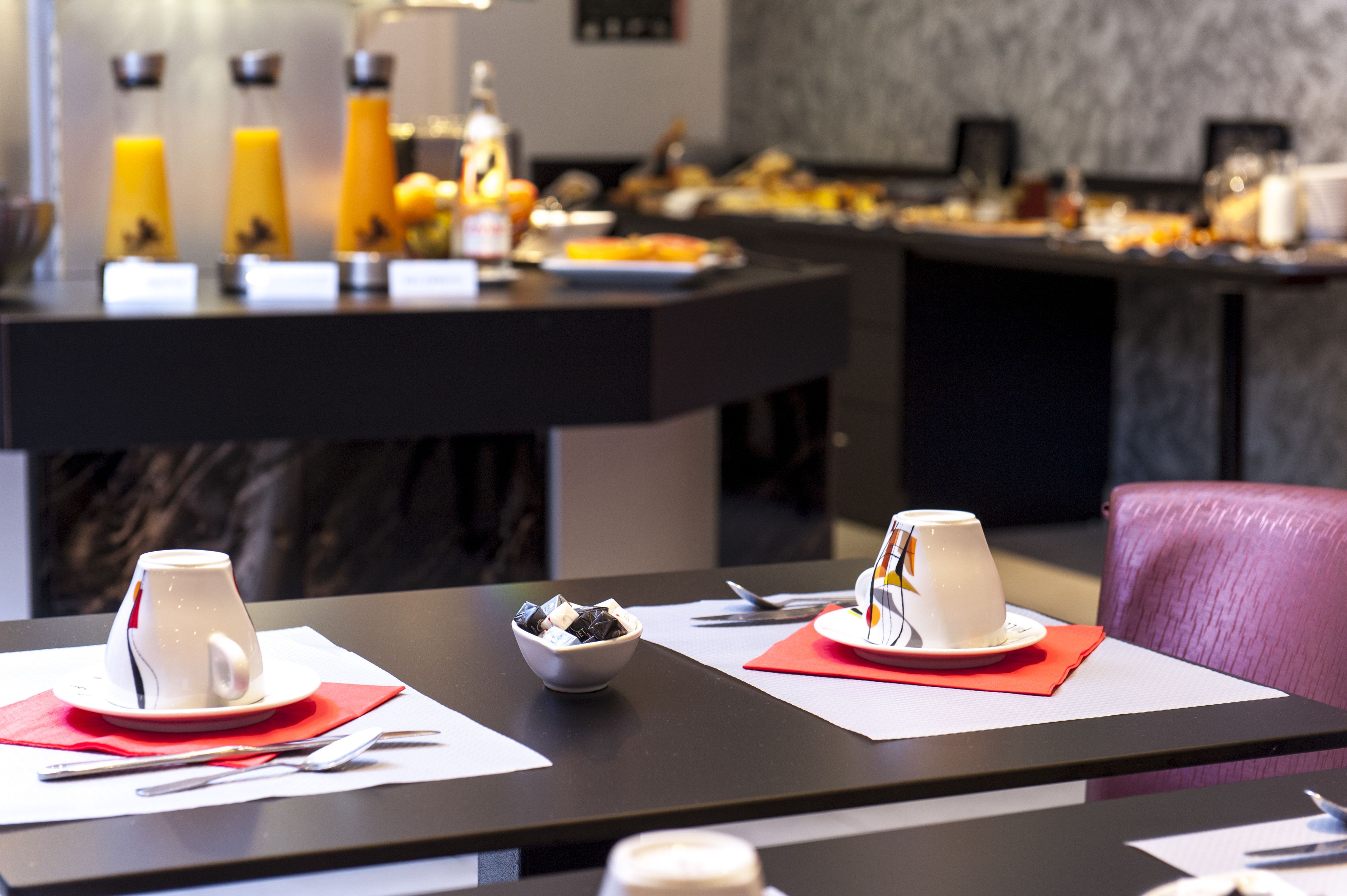 Une table dressée pour le petit-déjeuner avec deux tasses blanches posées sur des serviettes rouges, un saladier de fruits et un buffet en arrière-plan.