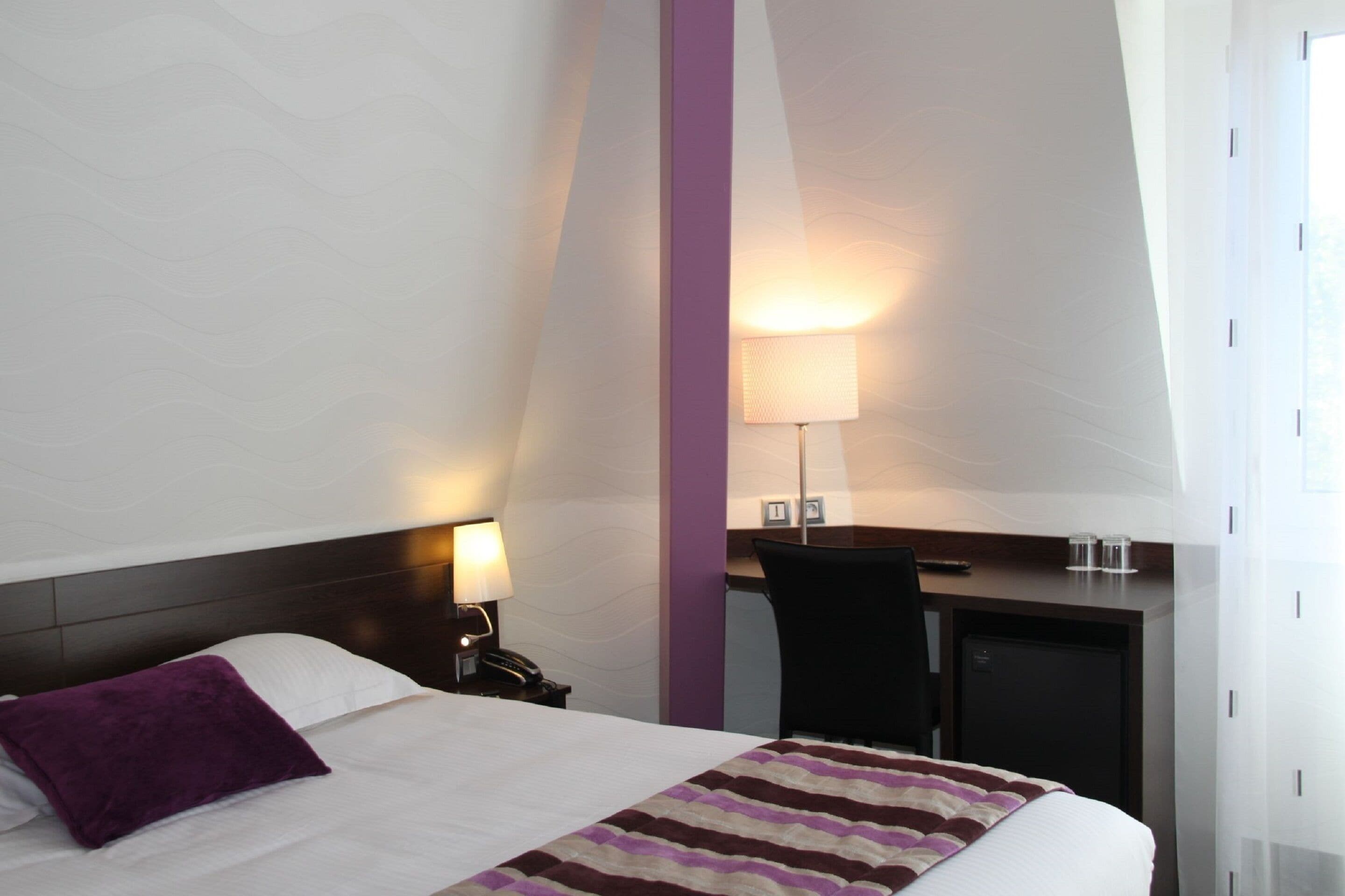 A hotel room with a bed, a desk, a chair, a lamp, and a nightstand.