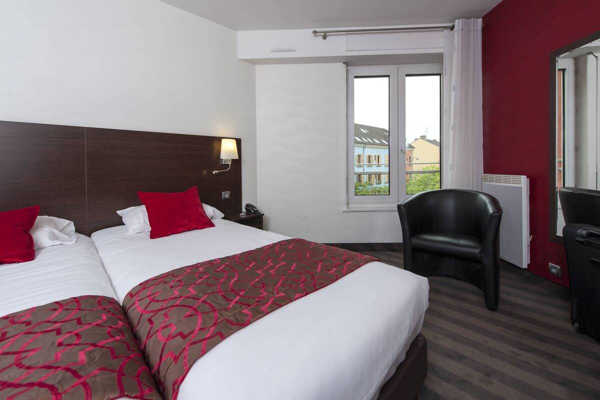Habitación de hotel con dos camas, un escritorio, una silla y ventana con vista a edificios.
