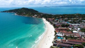 Vista aérea de una zona costera con playa, una pequeña isla y una área residencial con casas y piscina.