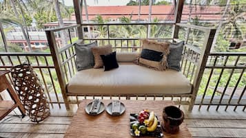 A wooden deck with a cushioned seating area, a woven chair, and a table with fruit and a clay pot.