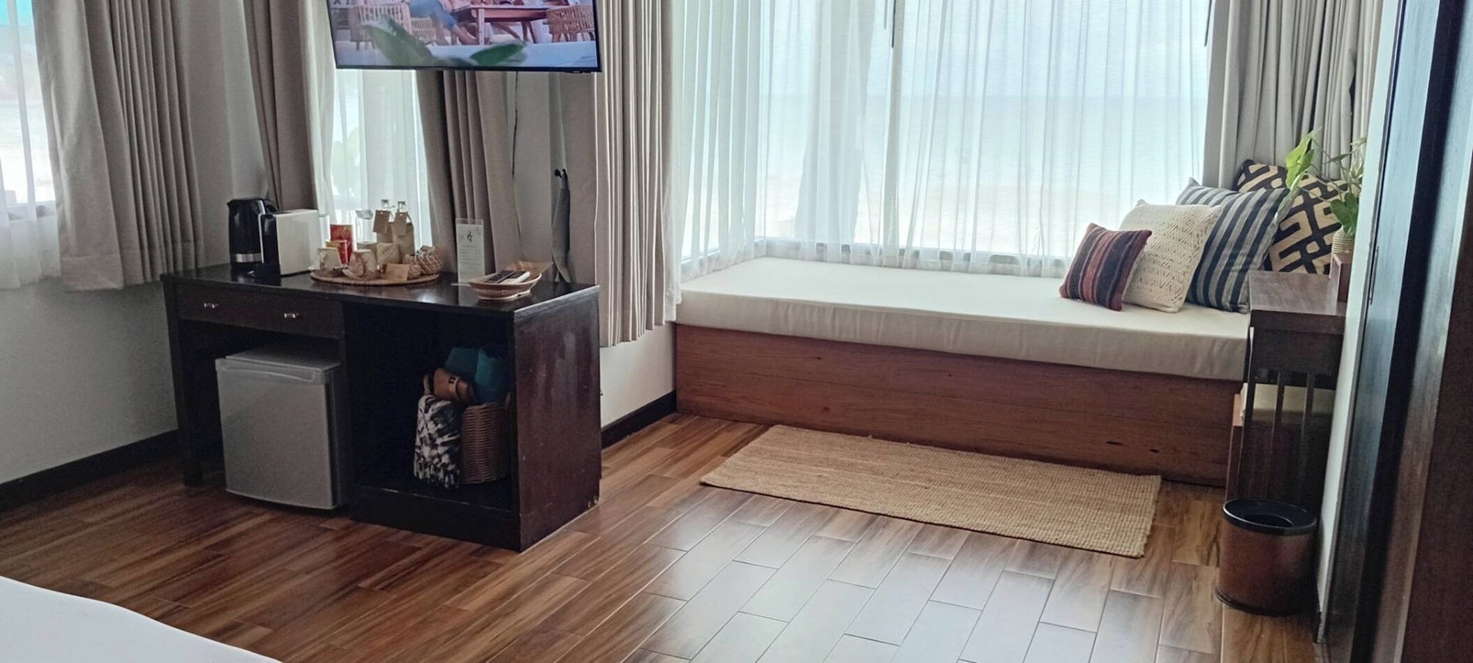 Una habitación con piso de madera, un escritorio pequeño con cafetera, un televisor, un banco acolchado con almohadas y una ventana con cortinas translúcidas.