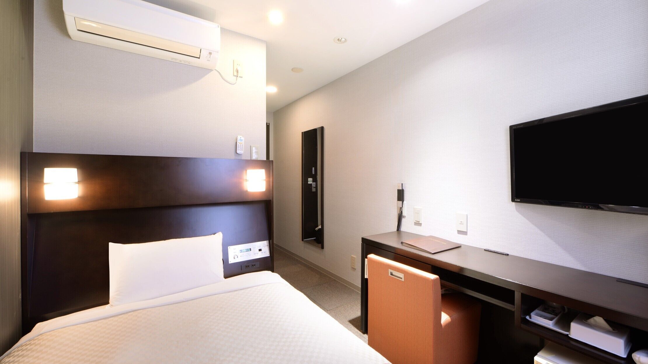 A hotel room with a bed, bedside table, TV, and a wall-mounted air conditioner.