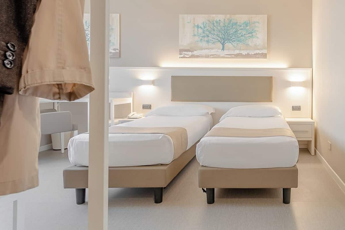 A hotel room with two beds, a headboard with integrated lighting, a painting on the wall, and a coat rack.