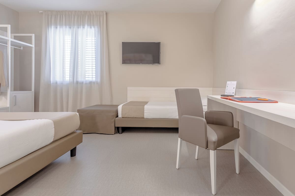 A hotel room with a bed, a desk with a chair, a small table, a TV, and a window with curtains.