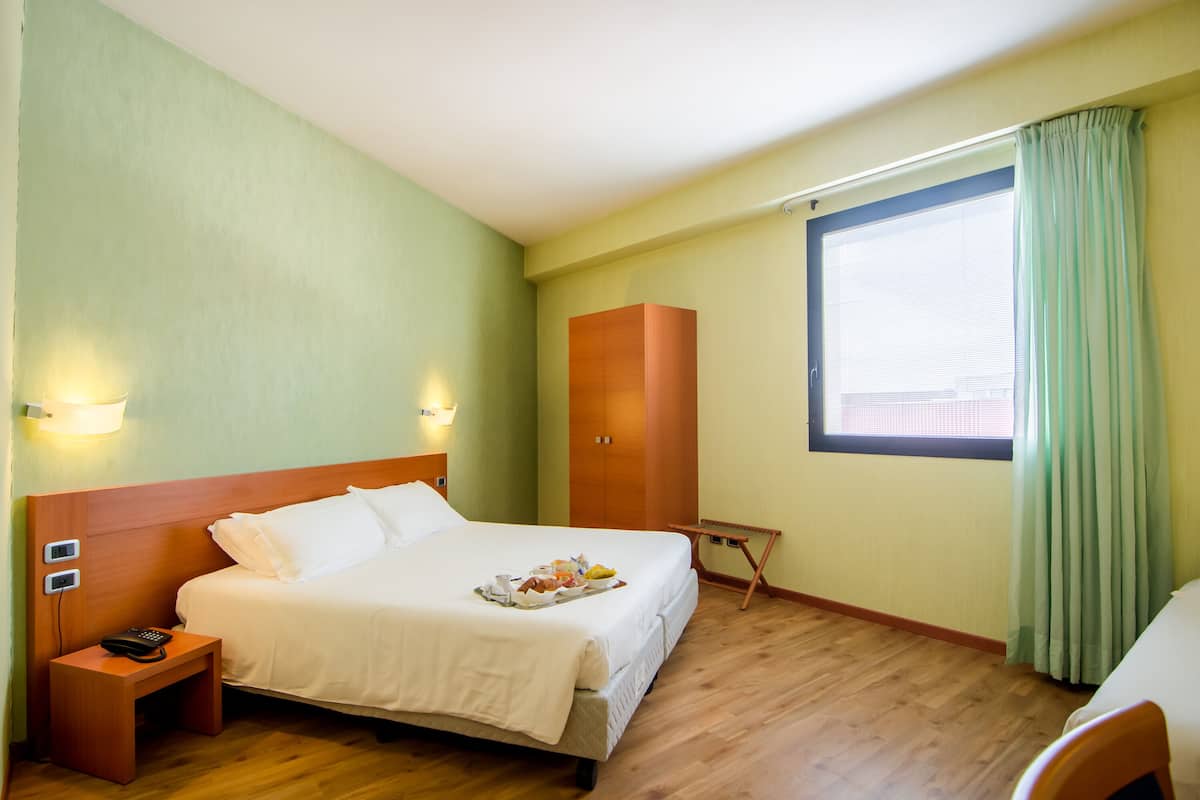 A hotel room with a bed, bedside table, lamp, wardrobe, and a window with curtains.