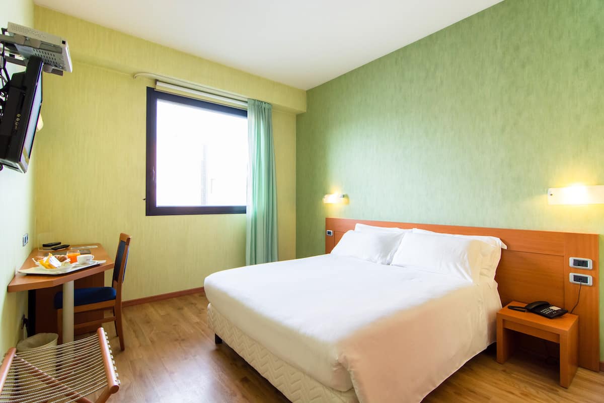 A hotel room with a bed, bedside table, chair, and a small table with breakfast items.