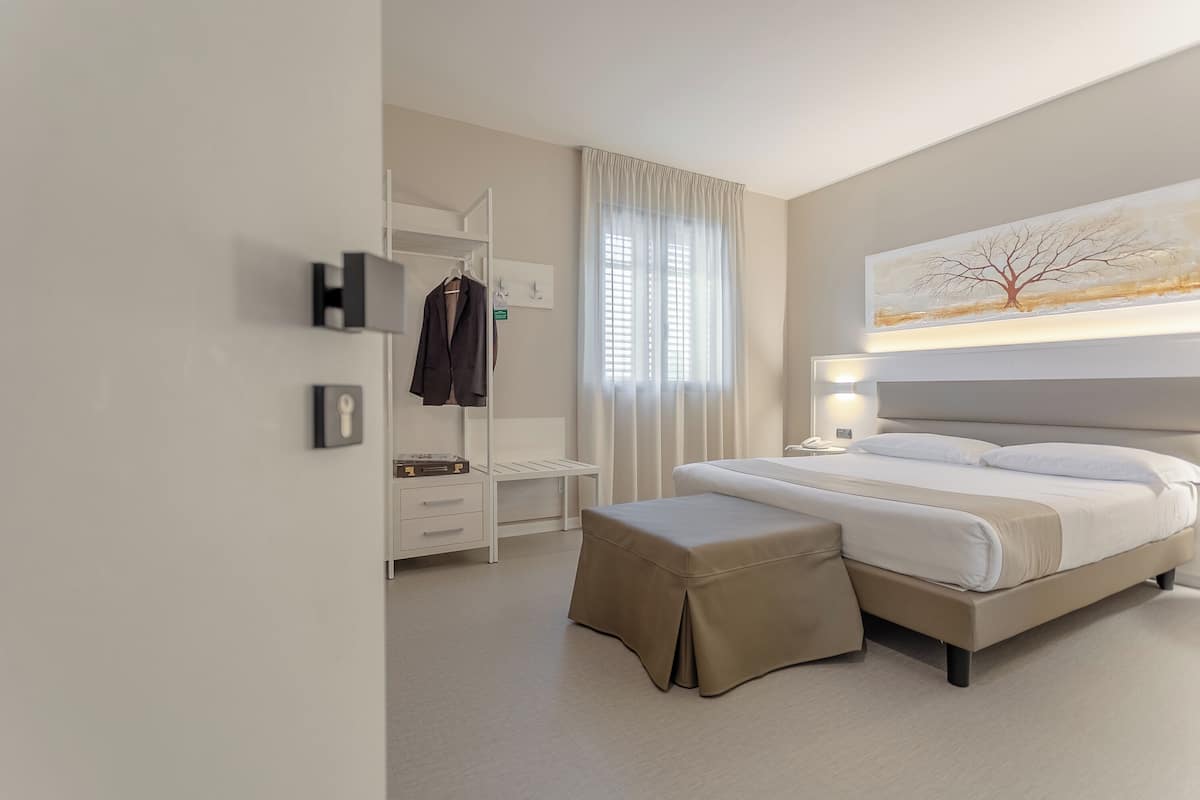 A modern hotel room with a large bed, a bedside table, a wardrobe, and a window with curtains.