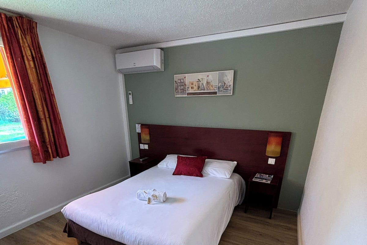 A neatly made bed with a brown headboard, a red pillow, and a white blanket. There is a small table beside the bed with a book and a bottle. The room has light green walls and wooden flooring.