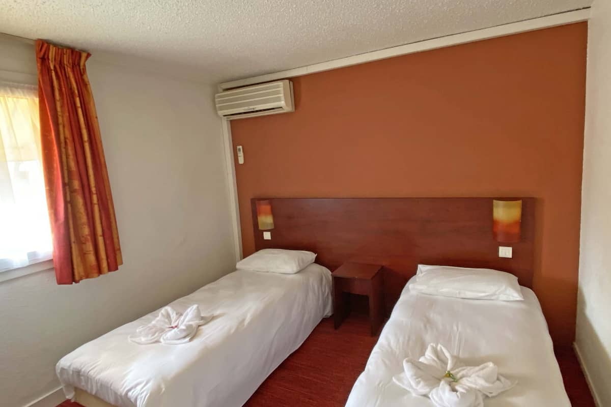 A hotel room with two single beds, a wooden headboard, an air conditioning unit, and a window with curtains.