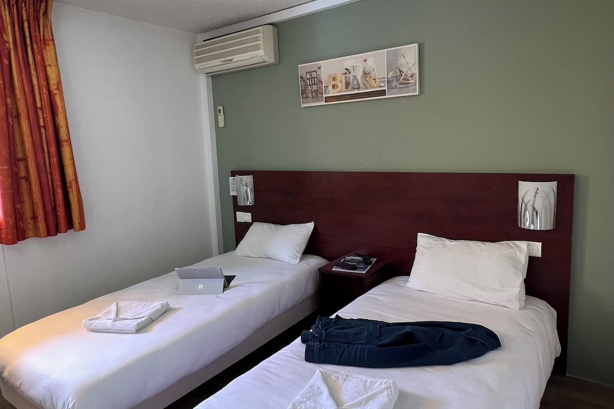 A hotel room with two single beds, a nightstand with a phone and a lamp, a wall-mounted air conditioner, and a framed picture above the beds.