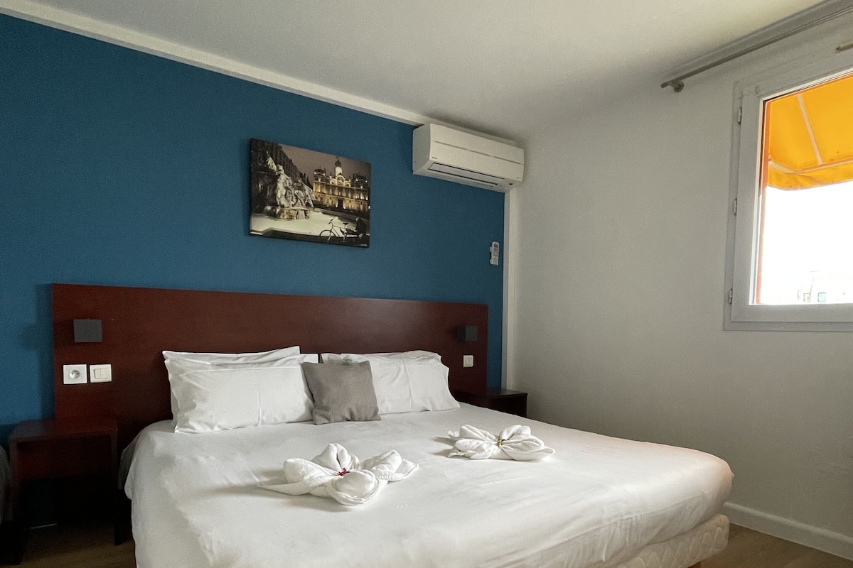 A neatly made bed with white linens and towels arranged in a bow shape, a wooden headboard, a nightstand, a wall-mounted air conditioner, and a window with a yellow curtain.