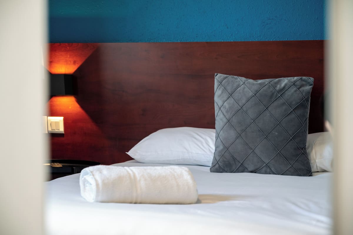 A neatly made bed with a white pillow and a grey quilted pillow, a white towel, and a wall-mounted light fixture.