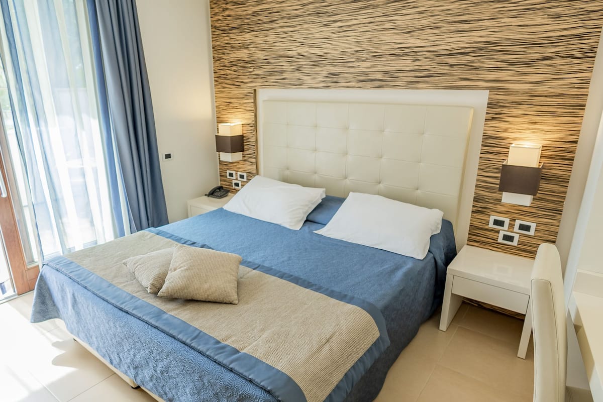 A modern bedroom with a large bed, a nightstand, a chair, and a view of the outdoors.